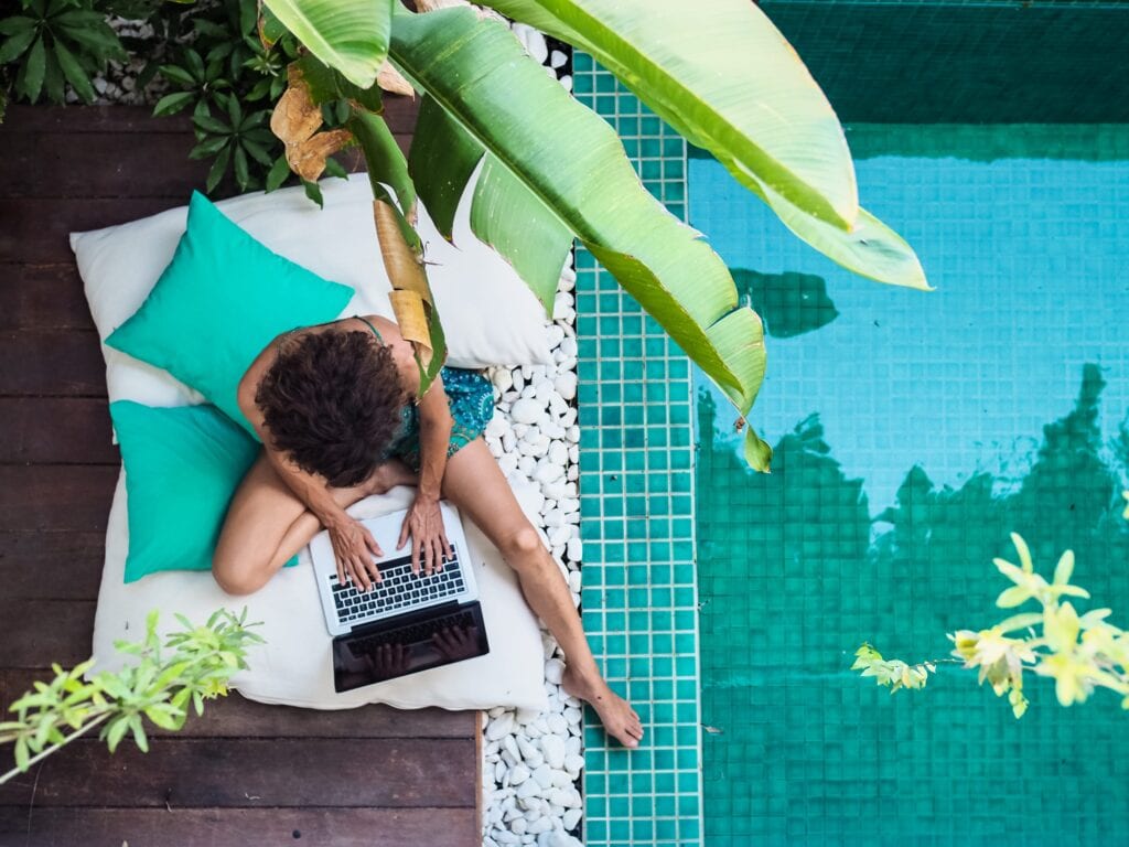 Eine Frau sitzt neben dem Pool und arbeitet am Laptop. -New Work